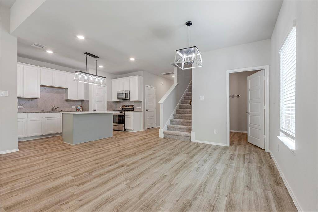 This modern open-concept space features a kitchen with white cabinetry, an island, and stainless steel appliances. It has wood-look flooring, recessed lighting, and a central staircase, creating a bright and inviting atmosphere.