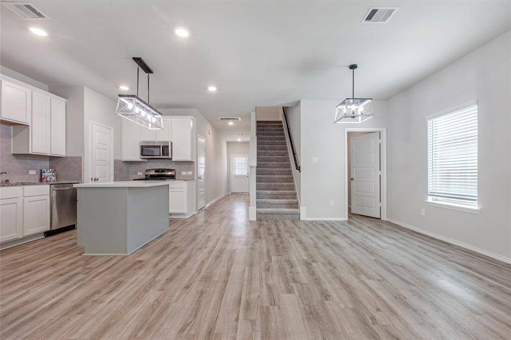 This photo showcases a modern open-concept kitchen and living area. It features light wood flooring, white cabinetry, stainless steel appliances, and contemporary light fixtures. A staircase leads to the upper level, and large windows provide ample natural light.