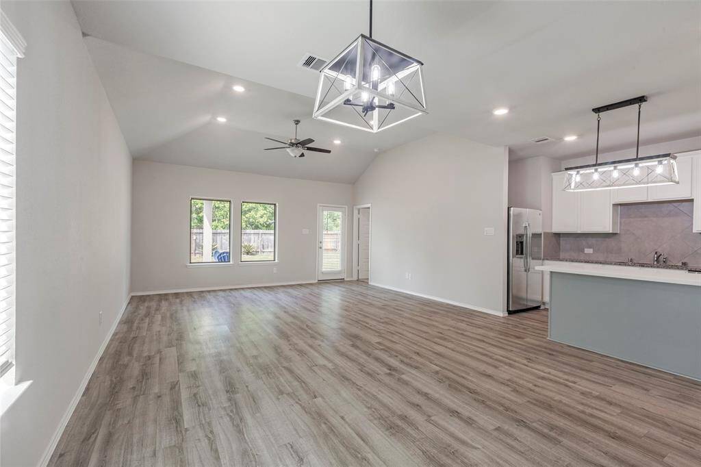 This is a spacious open-plan living area with high ceilings, modern light fixtures, and wood-style flooring. It features large windows for natural light, a ceiling fan, and a contemporary kitchen with white cabinetry and stainless steel appliances.
