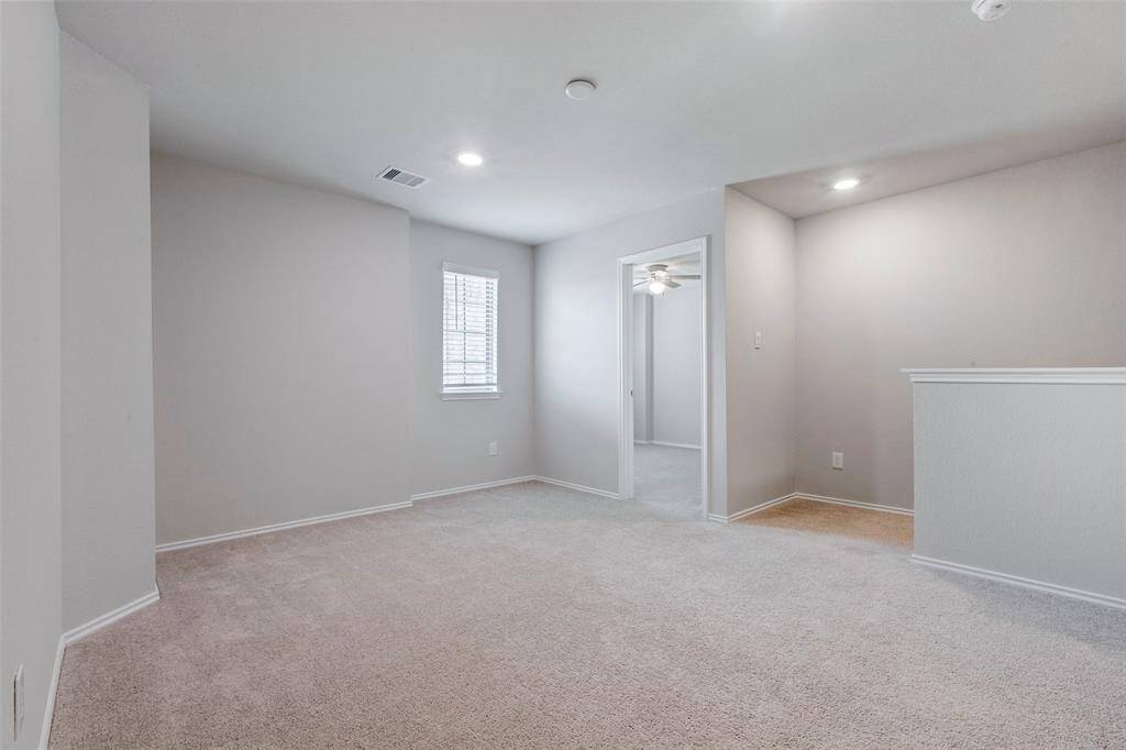 This image shows a bright, carpeted room with neutral walls, recessed lighting, and a small window. There's an open doorway leading into another space with a ceiling fan. Ideal for a cozy living area or office.