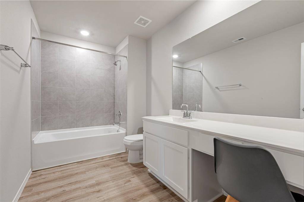 This bathroom features a clean, modern design with neutral tones. It includes a bathtub with a tiled shower, a large mirror above a sleek white vanity, and wood-style flooring, creating a bright and inviting space.