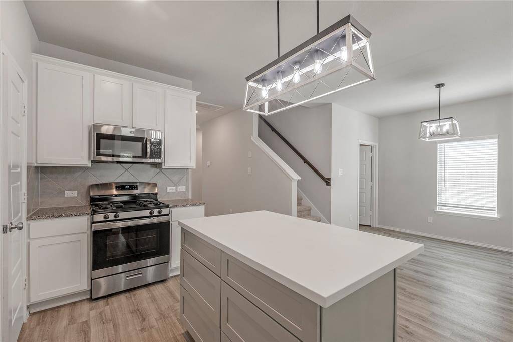 Modern kitchen featuring a sleek island with pendant lighting, stainless steel appliances, white cabinetry, and wood-look flooring. Adjacent dining area and staircase add to the open, bright layout.