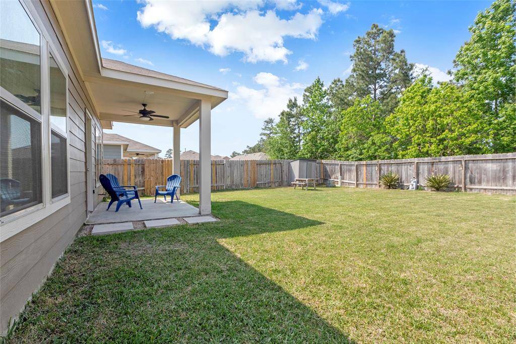 Spacious backyard with a covered patio featuring seating and a ceiling fan. The yard is fenced, grassy, and includes a small shed and trees for added privacy. Ideal for outdoor relaxation and activities.