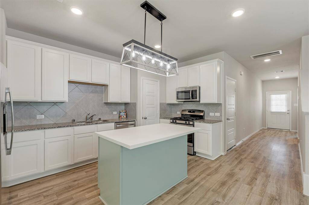 This modern kitchen features sleek white cabinetry, granite countertops, and stainless steel appliances, including a gas stove and microwave. The island with a light blue base offers additional workspace and seating. Recessed lighting and a central pendant light provide ample illumination, while wood-style flooring adds warmth to the space.