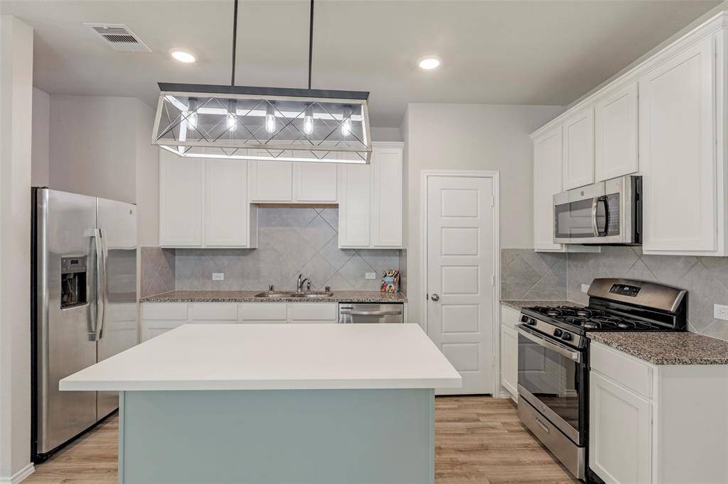 This modern kitchen features stainless steel appliances, a large island with a sleek light fixture above, white cabinetry, granite countertops, and a gas stove, offering both functionality and style.