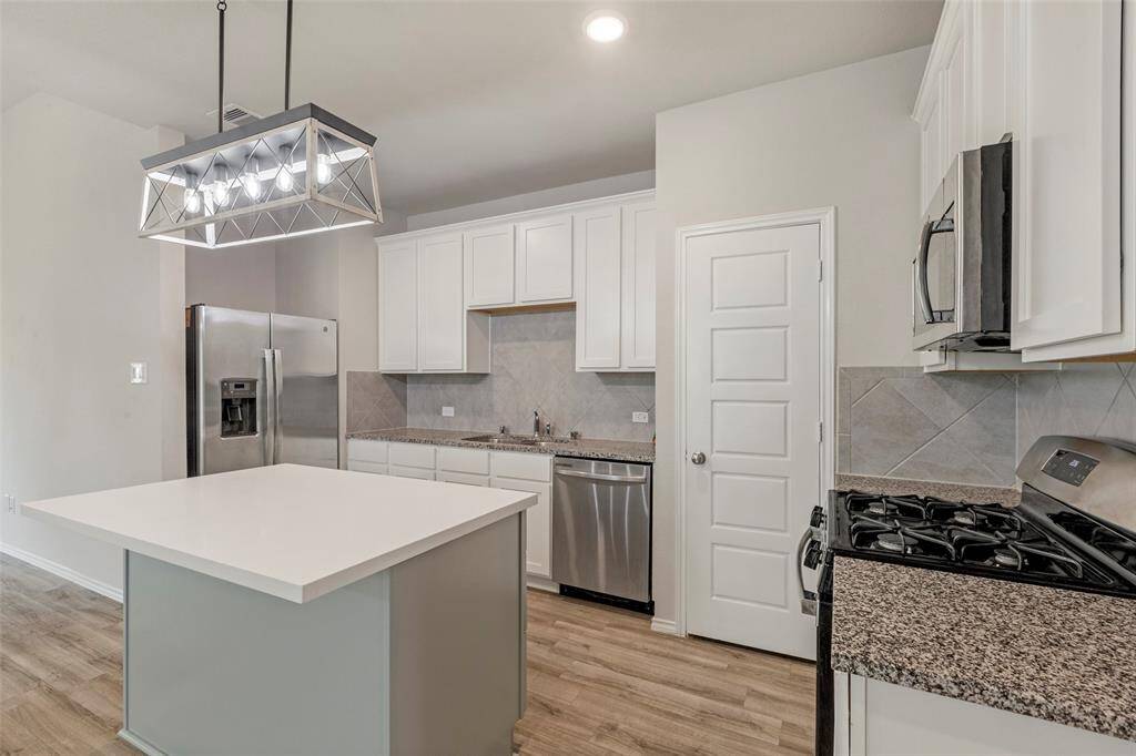 This modern kitchen features sleek white cabinetry, granite countertops, and stainless steel appliances, including a fridge and gas stove. The large island provides ample workspace, and the stylish light fixture adds a contemporary touch, all set against a backdrop of light wood flooring.