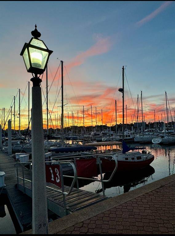 This photo captures a serene marina scene at sunset, featuring moored sailboats, a glowing lantern, and a vibrant sky with orange and pink hues, offering a picturesque seaside view.