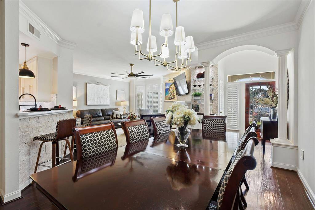 This photo showcases a stylish open-plan living and dining area with elegant features. It includes a spacious wooden dining table with patterned chairs, a modern chandelier, and a spacious living space with a mounted TV and fireplace. The room is well-lit with natural light from large windows and a glass door leading outside.