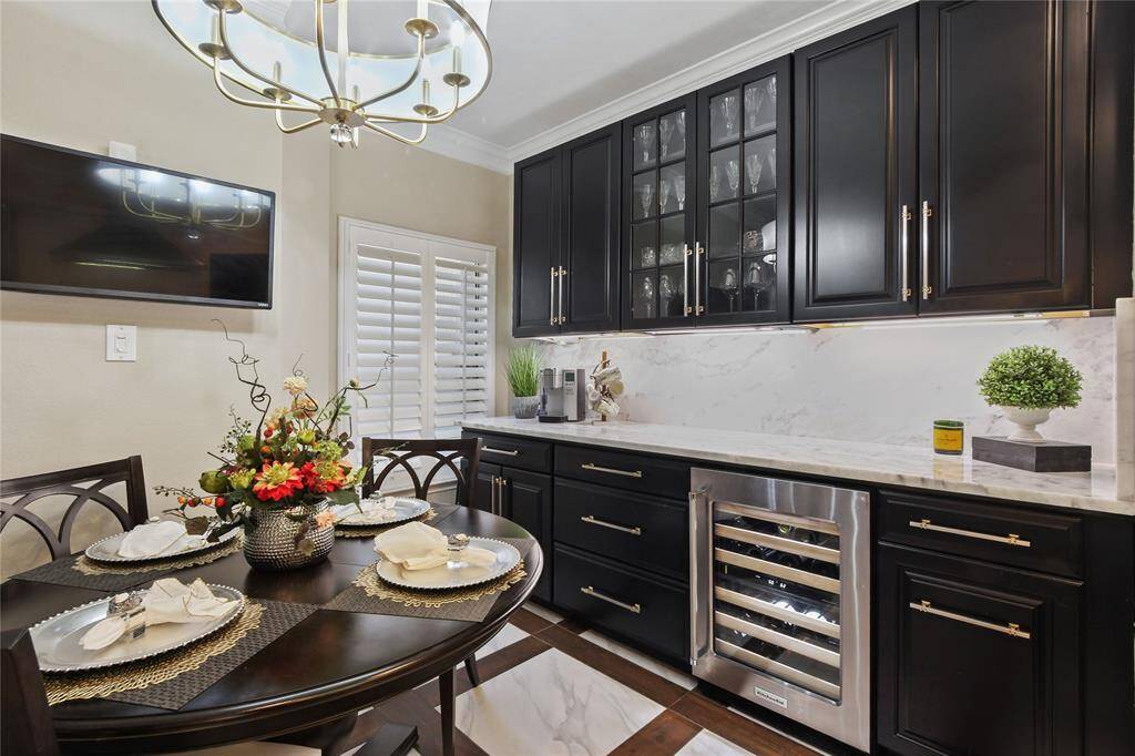 This elegant kitchen features sleek dark wood and white cabinetry with glass accents, a wine cooler, and a marble backsplash. A round dining table is beautifully set, and a modern chandelier adds a touch of sophistication.