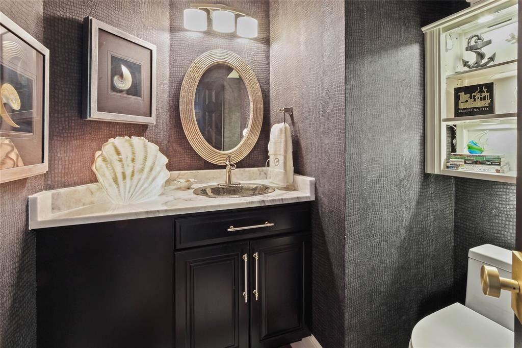 This is a stylish downstairs powder room featuring textured dark walls, a sleek vanity with a marble countertop, a decorative mirror, and elegant lighting. It includes creative art pieces and a small shelf with books, offering a modern, sophisticated look.