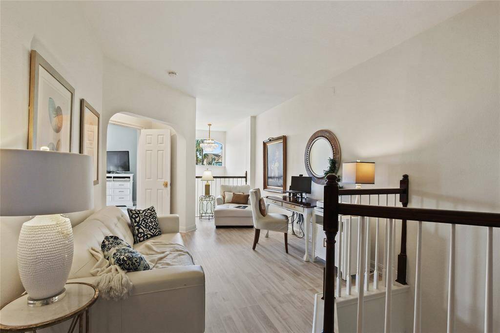 Bright, open upstairs home office/loft area with light wood flooring, cozy seating, and a workspace. Neutral decor and large windows enhance the inviting atmosphere. Ideal for relaxation and productivity.