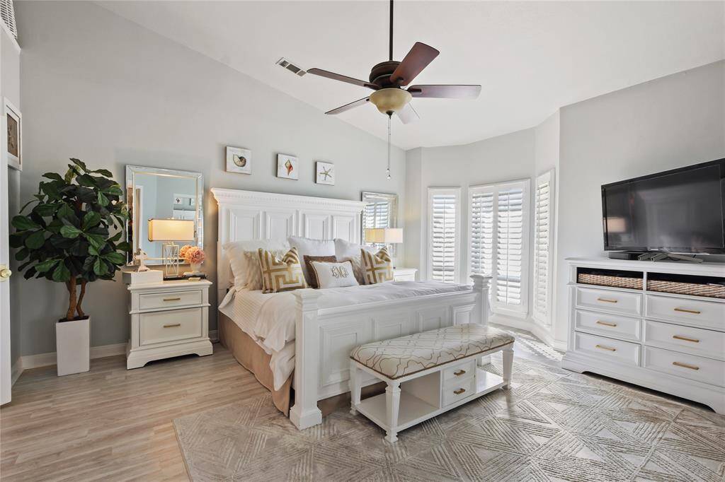 This bright, spacious one of Primary bedrooms... features a vaulted ceiling, elegant white furniture, and a neutral color palette. It includes a large bed with stylish decor, a ceiling fan, and ample natural light from the bay window with plantation shutters. A cozy seating area and modern entertainment setup complete the space.