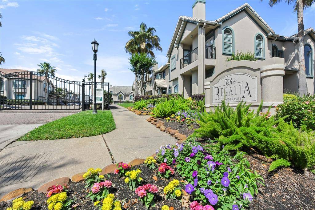 This photo showcases a gated townhome community entrance with well-maintained landscaping, featuring colorful flowers and palm trees. The buildings have a mediterranean design with balconies and a welcoming atmosphere.