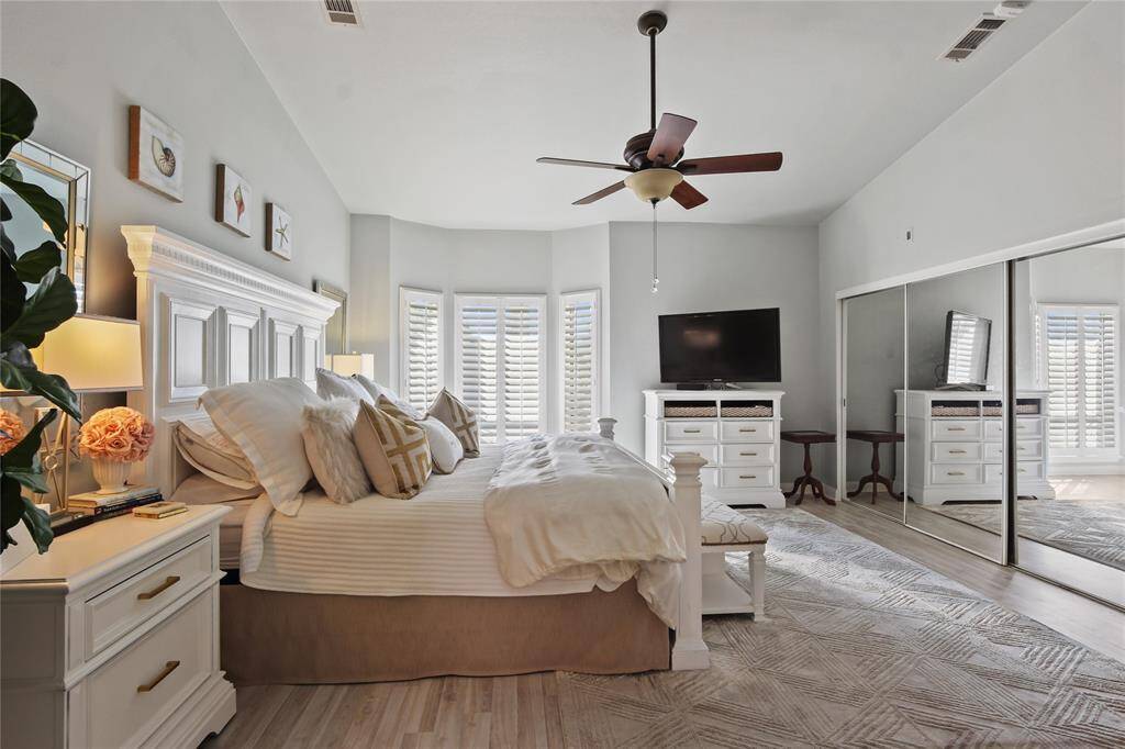This spacious bedroom features a cozy bed with elegant white furniture, a ceiling fan, and a large TV. Natural light streams through the bay windows with shutters, and mirrored closet doors enhance the room's brightness.