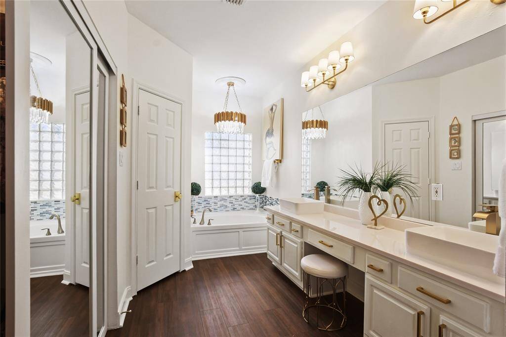 This elegant bathroom features a dual-sink vanity with gold accents, a spacious tub with stylish tile backsplash, and modern lighting fixtures. The room has a warm, inviting atmosphere with wood-look flooring and ample natural light.