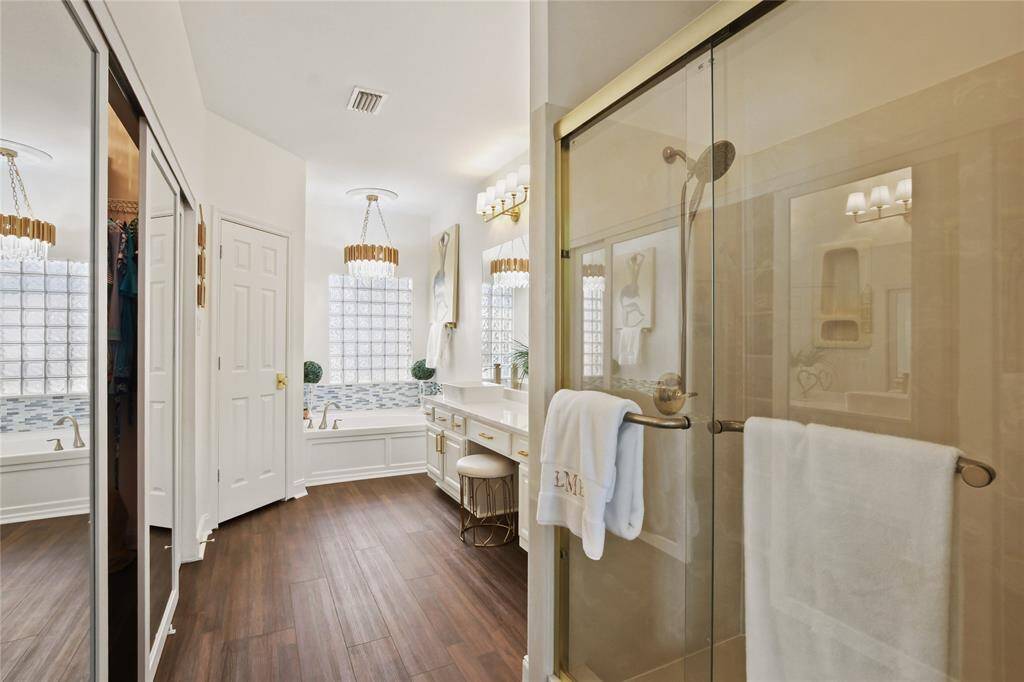 This bathroom features elegant finishes with a glass-enclosed shower, double vanity with ample lighting, and a luxurious soaking tub. The space is bright and inviting, complemented by wood-look flooring and modern fixtures.