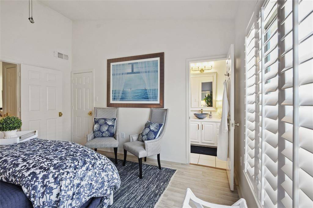 This bright and airy bedroom features large windows with shutters, allowing plenty of natural light. It has two cozy chairs, a stylish bedspread, and an en-suite bathroom with modern fixtures, creating a comfortable and inviting space.