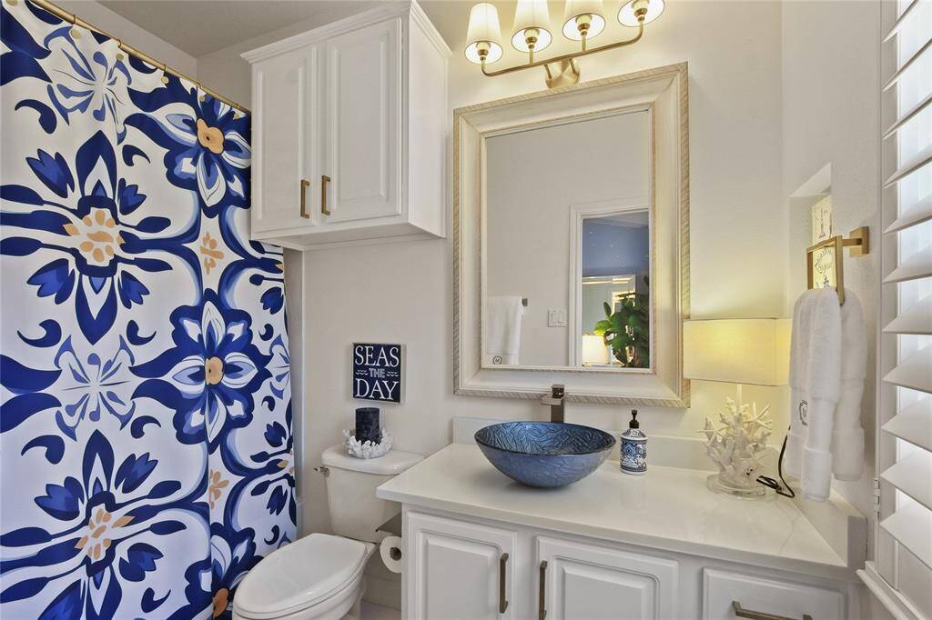 This bathroom features a stylish blue and white theme with a decorative floral shower curtain and a unique blue vessel sink. It includes a large framed mirror, overhead lighting, and white cabinetry for storage. A coastal vibe is accentuated by nautical decor and bright, natural light.
