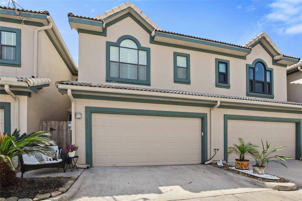 This is a two-story townhouse with a double garage, featuring a beige exterior.  The property includes a small front garden area with potted plants and a cozy seating spot, creating a welcoming entrance.