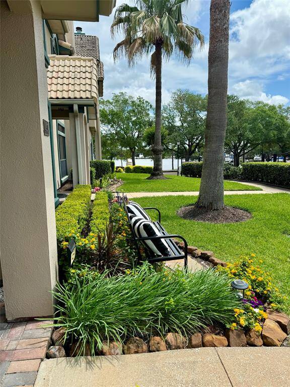 This photo shows a well-maintained front yard with lush landscaping, including a variety of plants and flowers. There's a pathway lined with hedges and a palm tree, leading towards a front yard scenic view of a lake. The exterior of the house features a tiled roof and stucco walls, giving it a charming, inviting appearance.