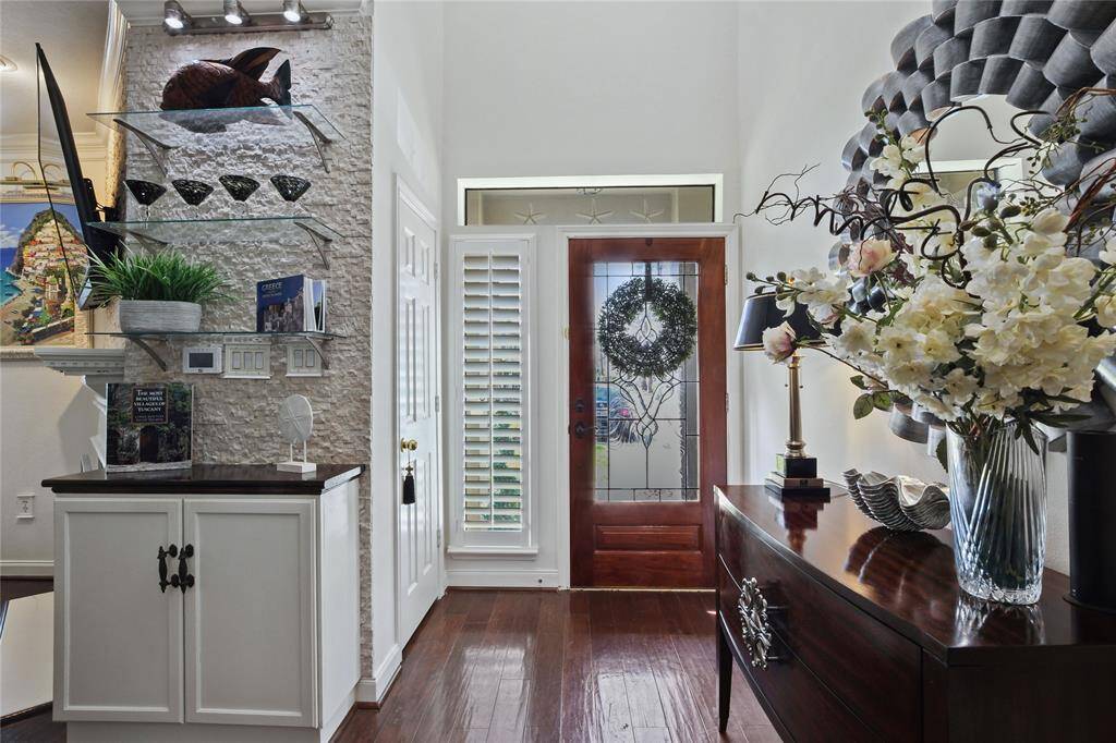This entryway features a welcoming wooden door with decorative glass, flanked by a window with plantation shutters. The space is elegantly styled with a dark wood console, floral arrangements, and minimalist decor, highlighted by a stone accent wall with glass shelving.