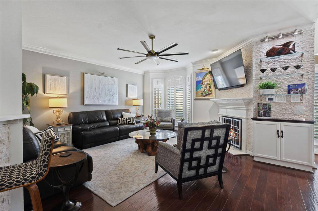 This inviting living room features hardwood floors, a cozy fireplace, and stylish decor. It offers a comfortable seating area with a leather sofa and modern chairs, complemented by a large TV and artistic accents. Natural light pours in through shuttered windows, enhancing the room's warm ambiance.