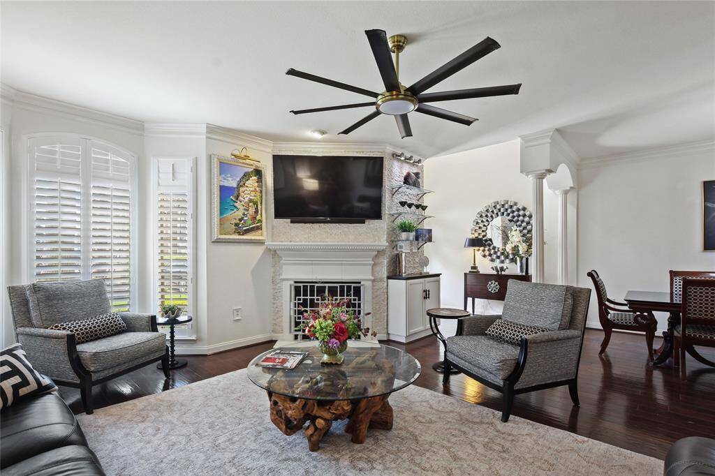 This inviting living room features elegant hardwood floors, a cozy fireplace with a mounted TV, and abundant natural light from large windows with shutters. The space is tastefully decorated with modern furniture, a unique coffee table, and a stylish ceiling fan, seamlessly connecting to a dining area.  Looks like a Model Home.