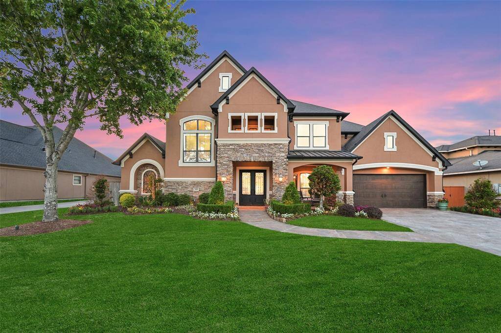 This stunning two-story stucco home features a modern design with a stone-accented facade and large windows. The well-maintained landscaping includes lush green grass and vibrant flower beds. A spacious lime coated driveway leads to a two-car garage, and the inviting entryway is framed by elegant architectural details and a spacious front patio.