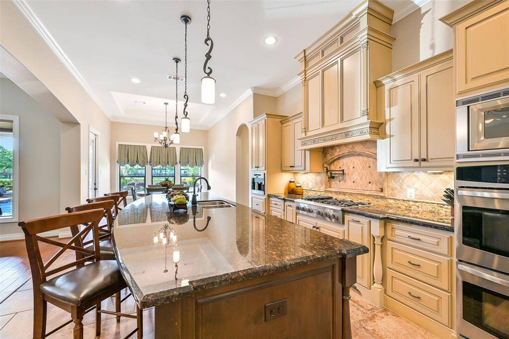 This bright, spacious kitchen features elegant cabinetry, granite countertops, and a large central island with seating. It includes stainless steel appliances, a stylish tile backsplash, and ample natural light from the windows, creating a welcoming atmosphere.