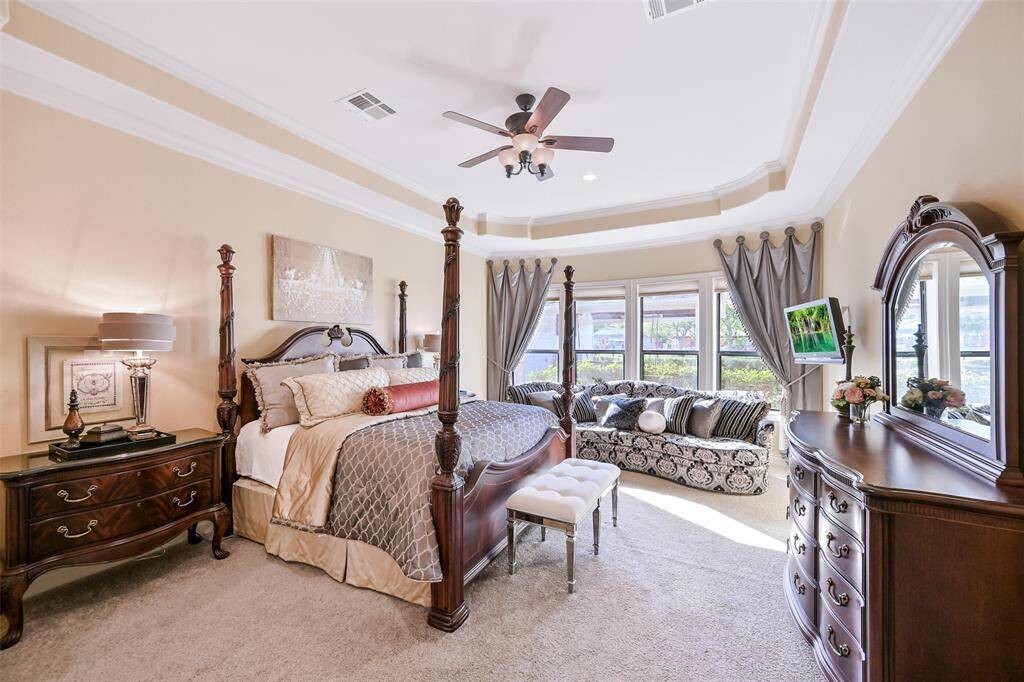 Spacious and elegant bedroom featuring tray ceilings, large windows with ample natural light, and elegant crown molding. Includes a cozy seating area, creating a luxurious and inviting atmosphere with views of the beautiful yard.