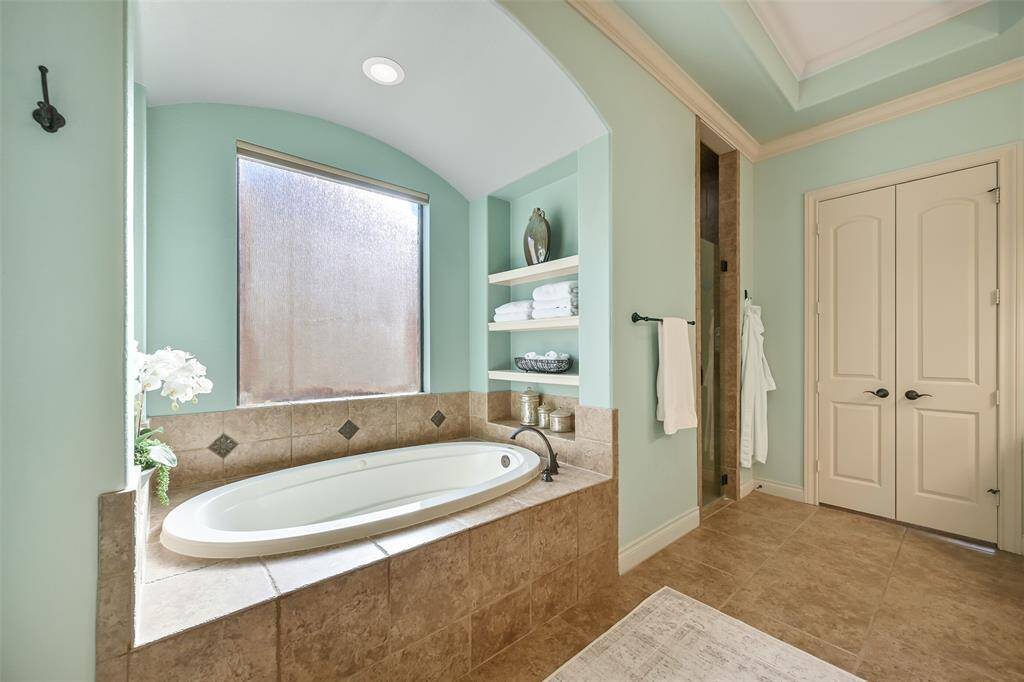 This bathroom features a spacious, elegant design with a large jacuzzi tub, built-in shelving with plenty storage. It includes a ceiling heat light, a separate shower area, and double doors, creating and serene spa-like atmosphere.