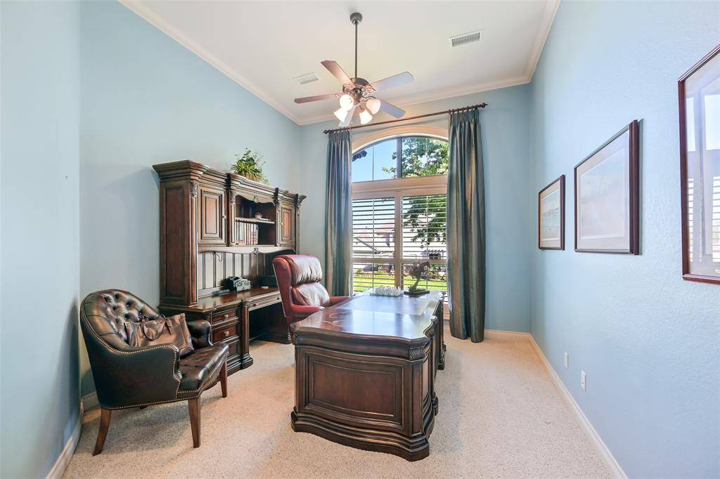 Step up into this bright home office with elegant crown molding and a lighted wall niche perfect for displaying art. The room features high ceilings, blue walls, and a large window with plantation shutters, offering a view of the outside.