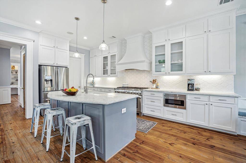 A sculptural vent hood, designer tile backsplash, and stainless steel appliances elevate the space, while a farmhouse sink and pendant lighting offer timeless touches.