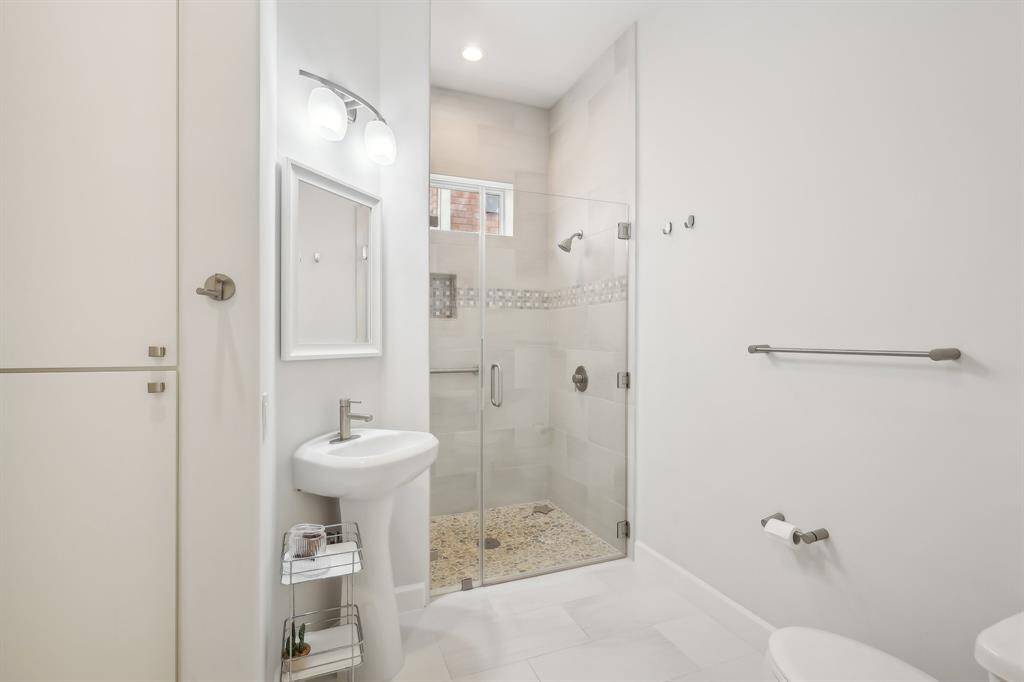 This first-floor full bathroom features a modern design with a glass-enclosed shower, a sleek pedestal sink, and neutral tile flooring. The space includes contemporary lighting, and convenient built-in cabinets for storage.