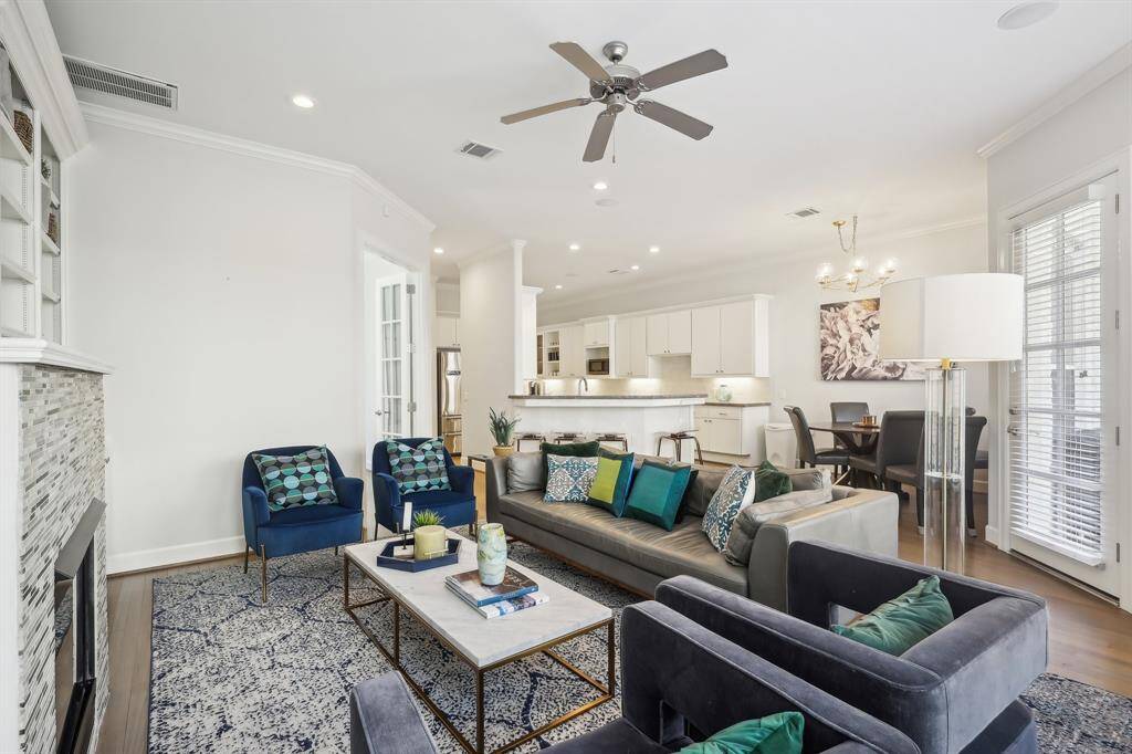 This photo features a bright, open-concept living area with a modern design. It includes a cozy seating arrangement with plush sofas and armchairs, a stylish fireplace, and an adjacent dining area. The kitchen is sleek with white cabinetry and a breakfast bar. Natural light floods the space through large windows, enhancing the airy feel.