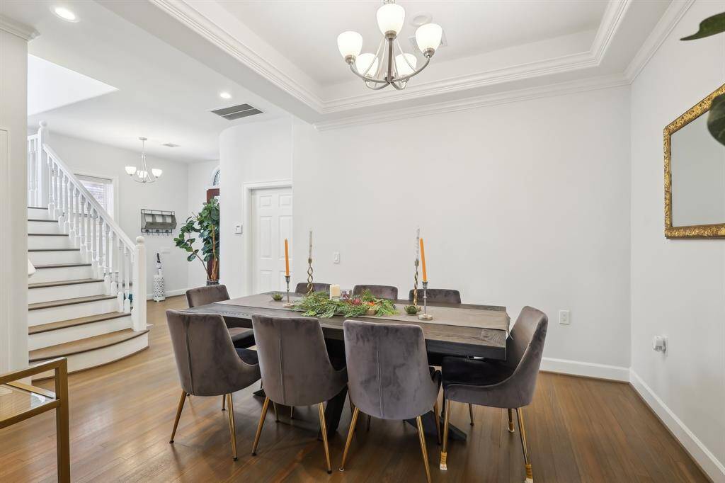 This photo showcases a bright and elegant dining area with a modern chandelier, a large wooden table, and plush chairs. The room features hardwood floors, a decorative ceiling, and is adjacent to a staircase, adding to the home's open and welcoming atmosphere.