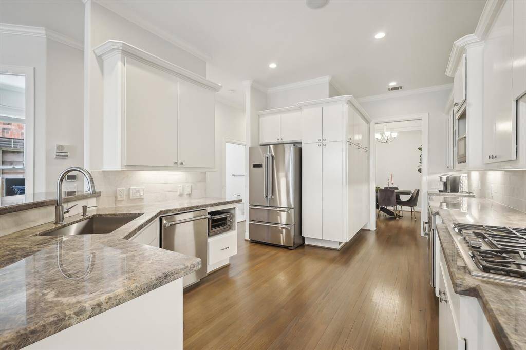 This kitchen features modern stainless steel appliances, sleek white cabinetry, and elegant granite countertops. The open layout with hardwood floors leads to a dining area, offering a bright and spacious cooking space.