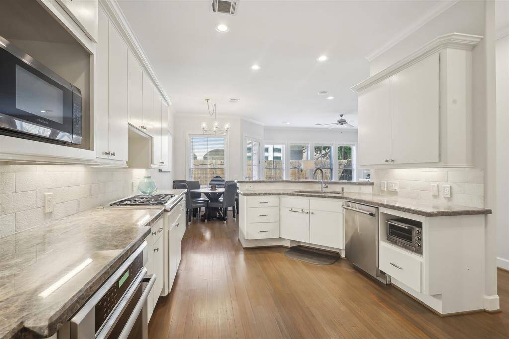 A bright, modern kitchen with white cabinetry, stainless steel appliances, and granite countertops. The open layout flows into a breakfast area with large windows and abundant natural light.