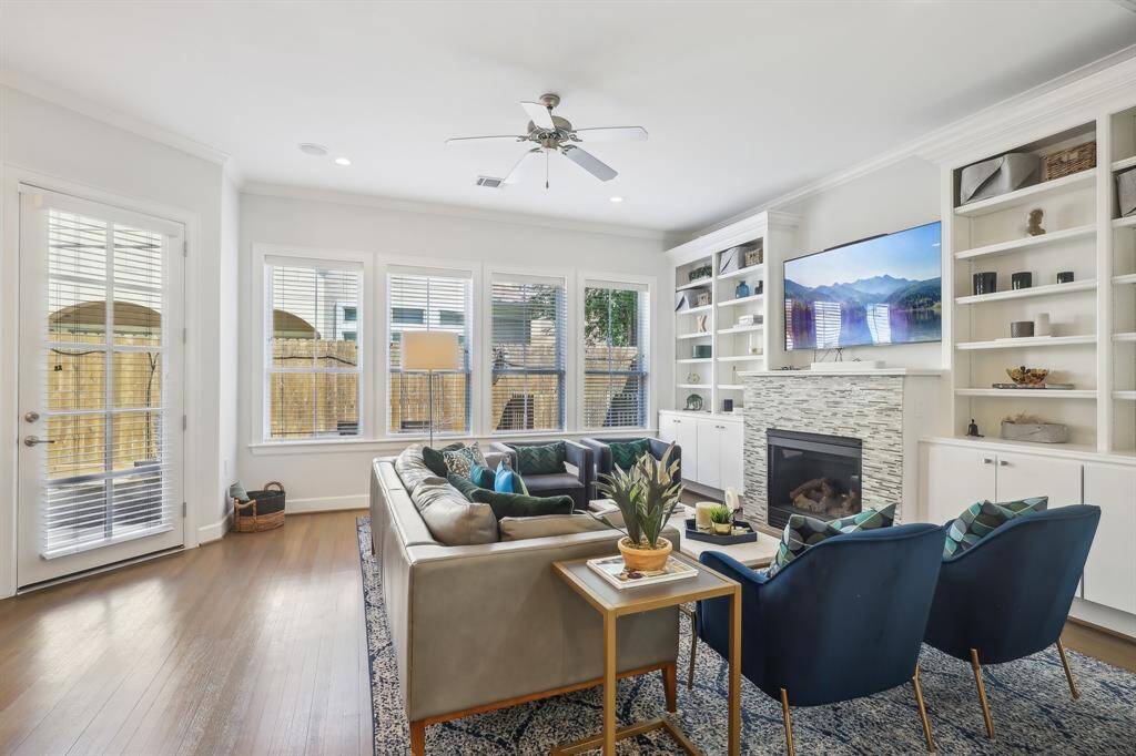 This bright living room features a cozy fireplace, built-in shelving, and a large sectional sofa. The space is enhanced by a wall-mounted TV, a ceiling fan, and ample natural light from multiple windows and a glass door leading to the outside.