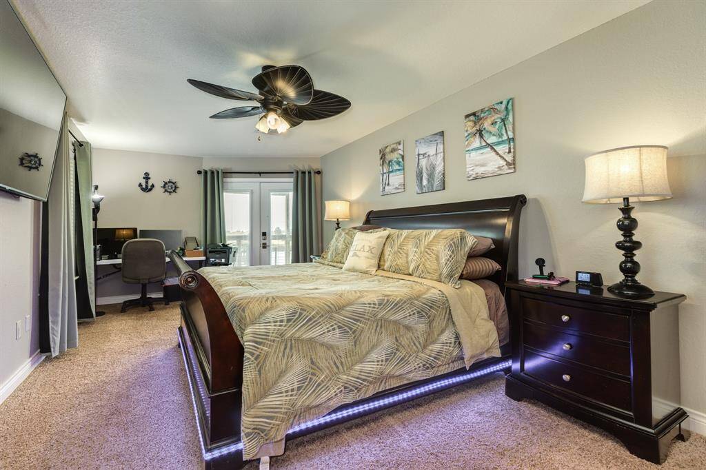 This inviting bedroom features a cozy carpeted floor, a stylish dark wood bed with under-lighting, and tropical-themed decor. A ceiling fan adds comfort, while a small office nook and French doors leading to deck enhances functionality and natural light.