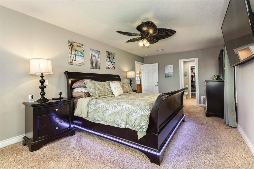 This spacious bedroom features a cozy carpeted floor, a stylish dark wood bed with under-bed lighting, and a ceiling fan. An en-suite bathroom and walk-in closet offer convenience, while wall art adds a touch of elegance.