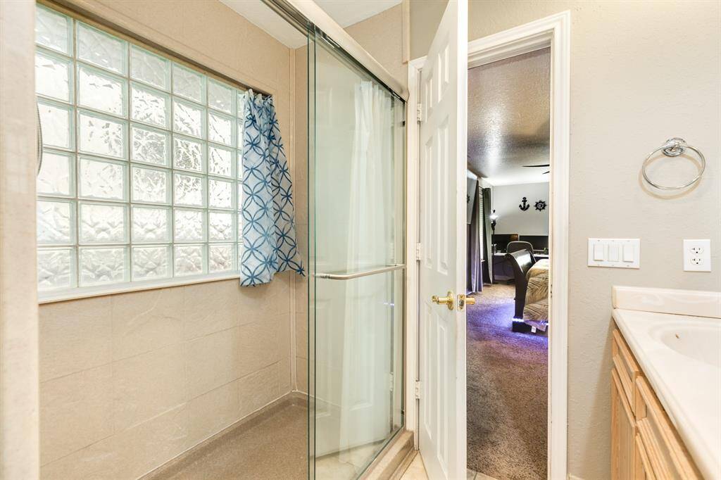 This photo features a bathroom with a glass shower, textured glass block window, and a view into a carpeted bedroom. The bathroom has light-colored walls and a countertop, creating a bright and inviting space.