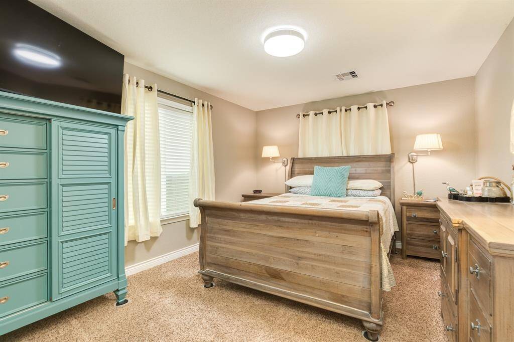 The 2nd bedroom with warm tones featuring a wooden bed, teal chest of drawers, and beige carpet. Bright natural light filters through light curtains, complemented by modern lighting fixtures and closet.