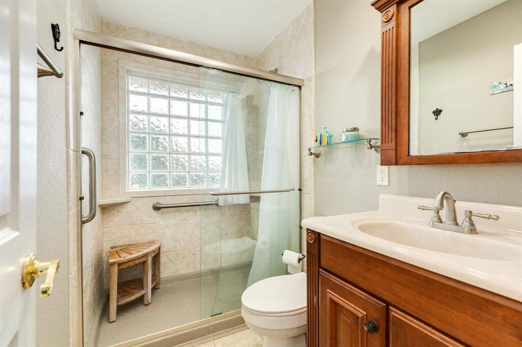 This bathroom features a glass-enclosed shower with a built-in bench and grab bars, a wooden vanity with a large mirror, and a frosted glass window for natural light.