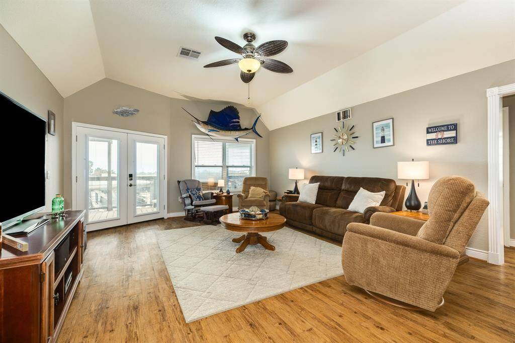 This inviting living room features vaulted ceilings, a ceiling fan, and ample natural light from large windows and French doors. The space is warmly decorated with a cozy sofa, recliner, and a nautical theme, including a mounted fish. The wood flooring adds a touch of elegance.