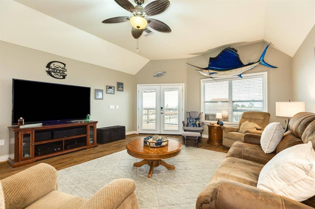 Spacious living room with vaulted ceiling, featuring cozy seating, large TV, ceiling fan, and natural light from french doors and windows. Decor includes nautical elements and wood flooring.