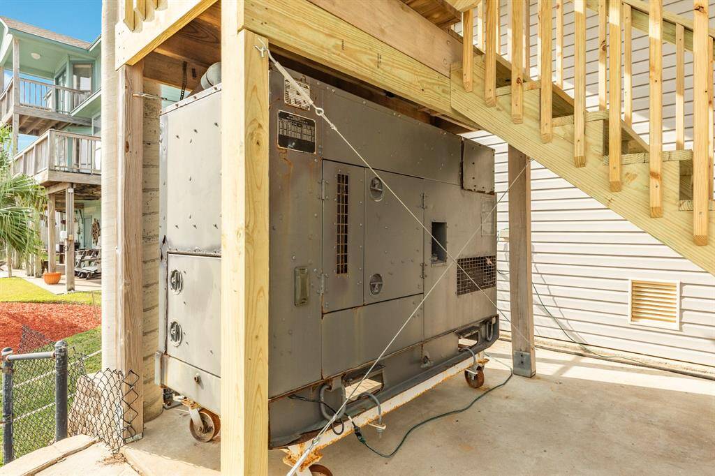 This photo shows an outdoor area with a large commercial generator set under a recently installed wooden staircase. The space is covered and part of a home with nearby siding and a fenced yard, suggesting additional utility and emergency power capabilities.