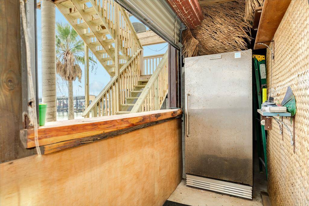 This photo features an outdoor area with a rustic vibe, including a metal refrigerator and wooden bar. A staircase leads to an upper level, and there's a view of a palm tree and waterfront in the background.