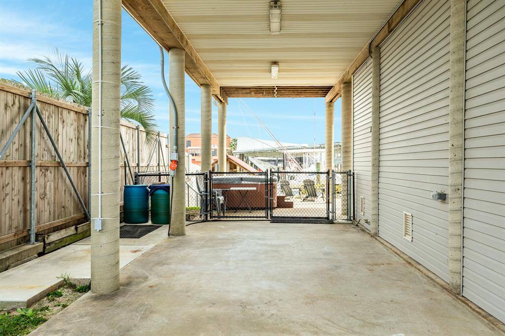 This photo shows a covered outdoor area with a concrete floor, surrounded by a wooden fence and metal gate. The space is supported by sturdy columns and features a view of a backyard with seating. It’s ideal for parking or outdoor activities.