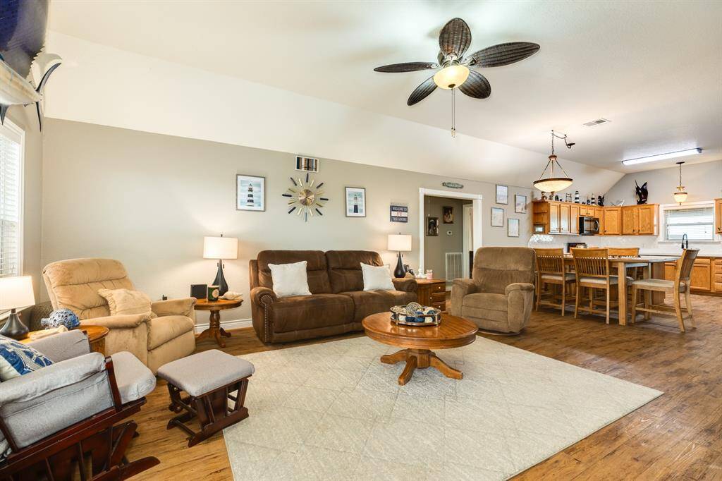 This inviting open-concept living area features a cozy seating arrangement with plush sofas and armchairs, a ceiling fan, and tasteful decor. The space seamlessly flows into a kitchen with warm wooden cabinetry and a breakfast bar, creating a perfect setting for family gatherings.